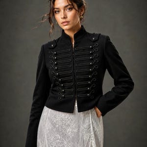 May include: A black military-style jacket with a high collar and decorative rope detailing. The jacket features multiple rows of small buttons and is worn over a white lace skirt. The jacket is a stylish piece of outerwear.