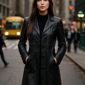May include: A long, black leather coat with a button-down front and a notched collar. The coat has pockets and is worn over a black top. The background shows a city street with a yellow bus and blurred figures.