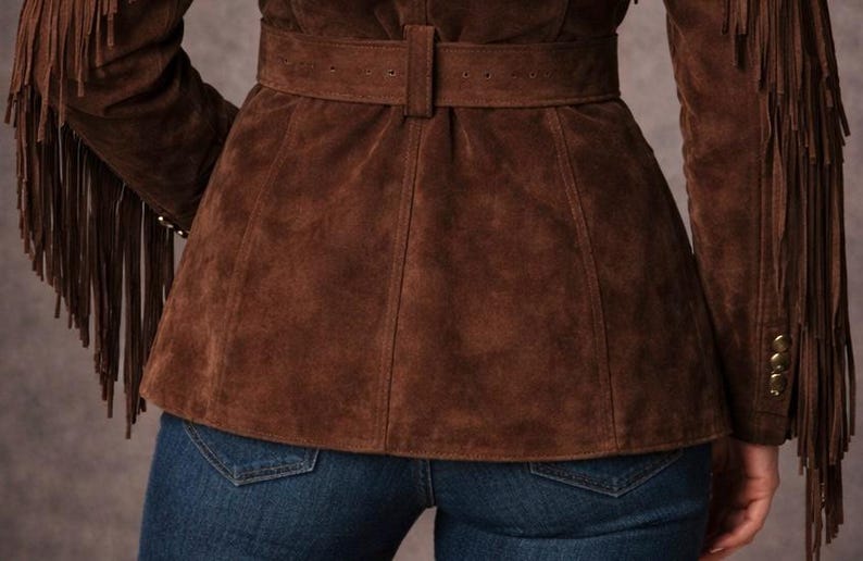 May include: A brown suede jacket with fringe detailing on the sleeves. The jacket features a belted waist and gold buttons. The model is wearing blue jeans. The jacket is a western-inspired style.
