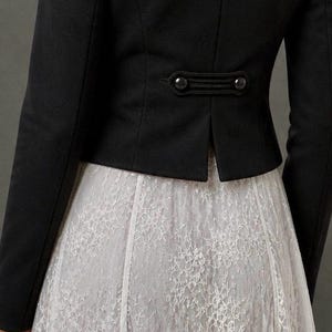 May include: A black jacket with long sleeves and decorative buttons at the back waist. The jacket is paired with a white lace skirt, creating a contrast in textures and colors. The overall style is elegant and classic.