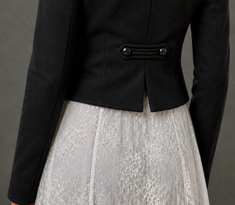 May include: A black jacket with long sleeves and decorative buttons at the back waist. The jacket is paired with a white lace skirt, creating a contrast in textures and colors. The overall style is elegant and classic.