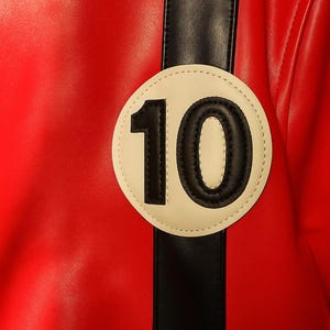 May include: Close-up of a red leather jacket with a black stripe and a white circular patch. The patch features the number 10 in black. The jacket has a smooth texture and a sporty design.