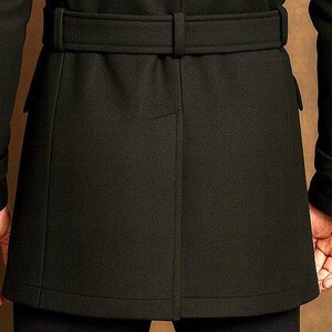 May include: A black coat featuring a belted waist with a matching fabric belt, long sleeves with buttoned cuff tabs, and a central vertical seam detail on the back. The coat has a structured fit and reaches mid-thigh length, showcasing clean lines.
