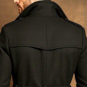 May include: A black coat featuring a high collar, shoulder epaulets, and a horizontal back yoke seam. The garment is constructed with structured tailoring, visible stitching along the seams, and a clean, solid black fabric finish throughout.