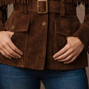 May include: A brown suede jacket with fringe detailing on the sleeves. The jacket features a belted waist with a gold-colored buckle, two front pockets, and button closures. The model is wearing blue jeans.