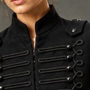 May include: Black military-style jacket with a high collar and decorative rope and button closures. The jacket features shoulder epaulettes with button details and a zipper closure. A stylish and unique outerwear piece.