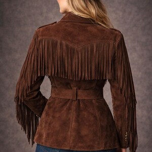 May include: Brown suede jacket with fringe detailing on the sleeves and back. The jacket features a belt at the waist and is paired with a matching brown cowboy hat. The model is wearing blue jeans. The jacket has gold buttons on the sleeves.