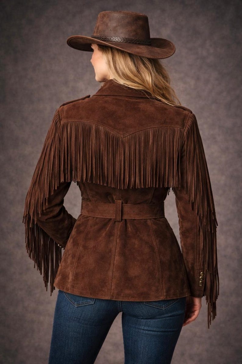 May include: Brown suede jacket with fringe detailing on the sleeves and back. The jacket features a belt at the waist and is paired with a matching brown cowboy hat. The model is wearing blue jeans. The jacket has gold buttons on the sleeves.
