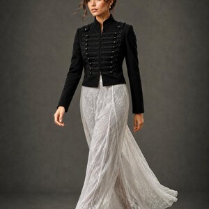 May include: A black military-style jacket with decorative silver buttons and a high collar. The jacket is paired with a flowing, white lace skirt. The outfit is completed with black high-heeled shoes, creating an elegant ensemble.
