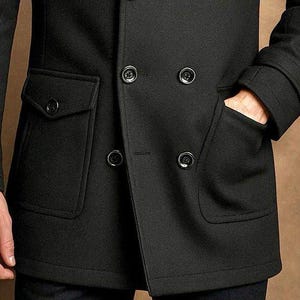 May include: A black double-breasted coat featuring a structured collar, long sleeves with buttoned cuffs, and two large front pockets with buttoned flaps. The coat is secured with four visible black buttons arranged in a double-breasted configuration.