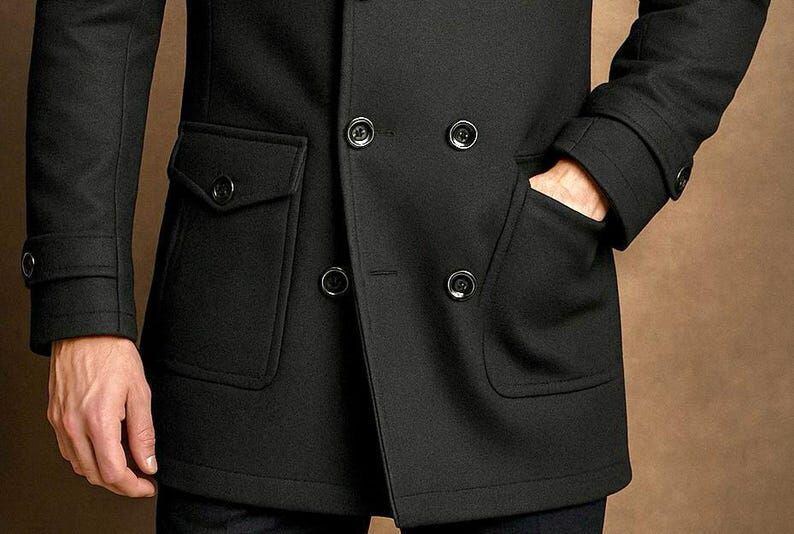 May include: A black double-breasted coat featuring a structured collar, long sleeves with buttoned cuffs, and two large front pockets with buttoned flaps. The coat is secured with four visible black buttons arranged in a double-breasted configuration.