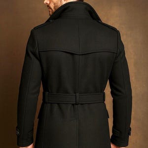 May include: A black trench coat featuring a wide collar, shoulder epaulets, and a matching waist belt with a buckle. The coat has long sleeves with buttoned cuffs, a back yoke, and a central vent at the hem, providing a structured silhouette.