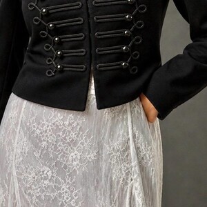 May include: A black military-style jacket with decorative buttons and rope detailing. The jacket is paired with a white lace skirt. The outfit is a blend of classic and romantic styles.