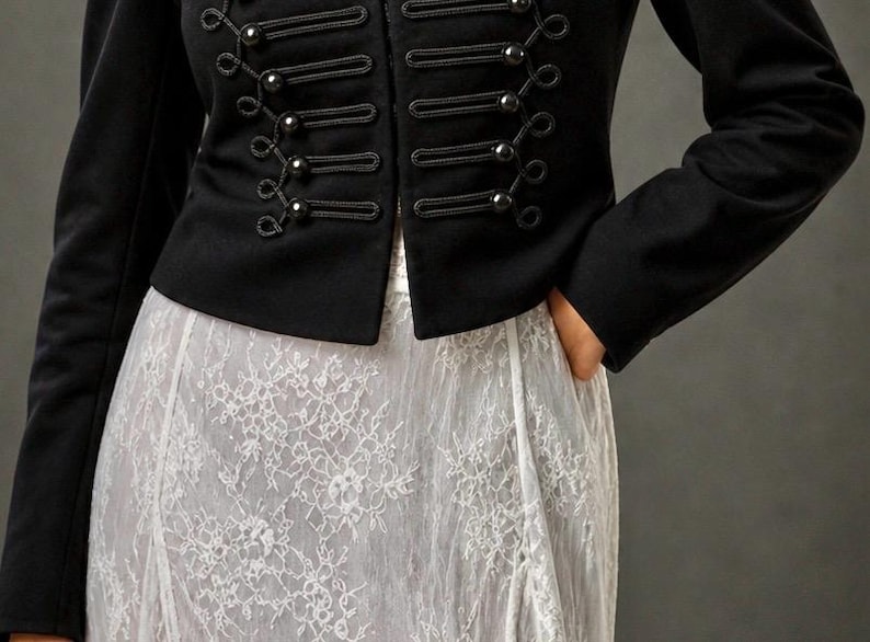 May include: A black military-style jacket with decorative buttons and rope detailing. The jacket is paired with a white lace skirt. The outfit is a blend of classic and romantic styles.