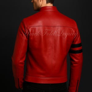 May include: A red leather jacket with black stripes on the sleeves and the text "Hawk Fold Legacy" on the back. The jacket has a stand-up collar and a zipper closure on the sleeves. The person is wearing black jeans.