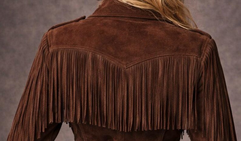 May include: A brown suede jacket with fringe detailing on the sleeves and back. The jacket has a classic western style with a collar and shoulder accents. The fringe adds a decorative element to the garment.