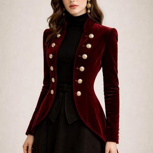 Puede incluir: Chaqueta de terciopelo burdeos con botones dorados. La chaqueta tiene un cuello alto y un corte entallado. La modelo lleva un cuello alto negro y una falda negra. Una prenda elegante.