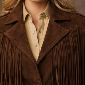 May include: A brown suede jacket with fringe detailing. The jacket has a collared neckline and button closures. A light tan button-down shirt is visible underneath the jacket.
