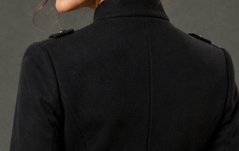 May include: Close-up of the back of a black coat. The coat has a high collar and two small decorative buttons on each shoulder. The fabric appears to be a smooth, woven material.