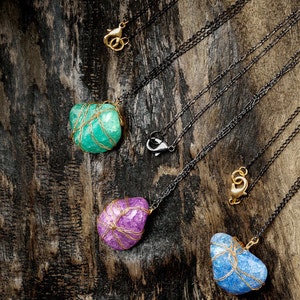 Puede incluir: Tres collares con colgantes de piedra de diferentes colores envueltos en alambre dorado. Las piedras son turquesa, p&uacute;rpura y azul. Los collares est&aacute;n sobre un fondo de madera oscura.