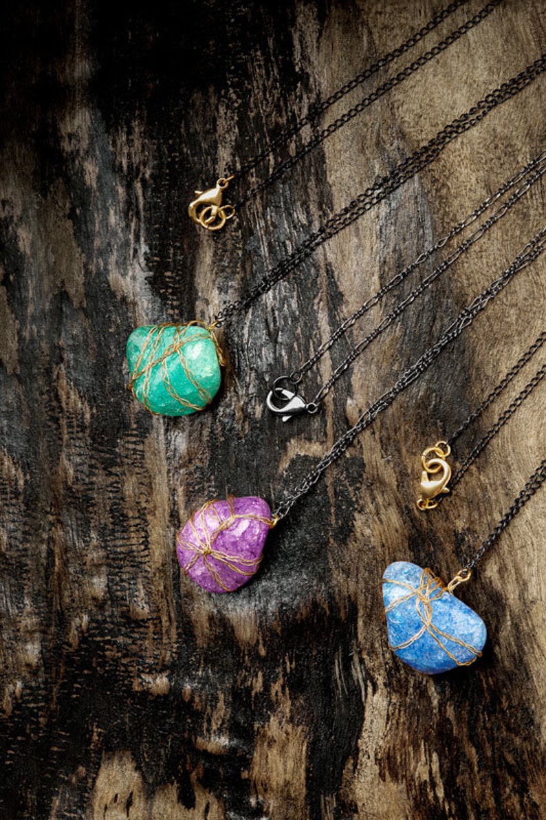 Puede incluir: Tres collares con colgantes de piedra de diferentes colores envueltos en alambre dorado. Las piedras son turquesa, p&uacute;rpura y azul. Los collares est&aacute;n sobre un fondo de madera oscura.