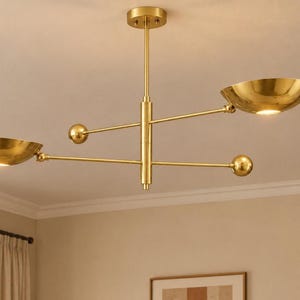 May include: A gold-toned metal chandelier featuring a central vertical rod with two horizontal arms extending in opposite directions. Each arm holds a bowl-shaped light fixture at one end and a decorative gold sphere at the other end.
