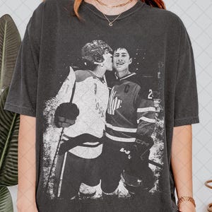 May include: Dark gray t-shirt featuring a black and white image of two hockey players. One player is kissing the other on the cheek. The players are wearing hockey uniforms and helmets. The shirt has a vintage, distressed look.