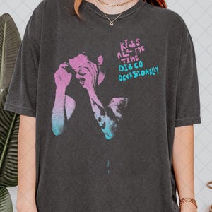 Puede incluir: Camiseta gris oscuro con un gráfico de una persona con acentos rosas y azules, con el texto "Kiss All The Time Disco Occasionally" en rosa y azul. La camiseta tiene mangas cortas y un corte holgado.
