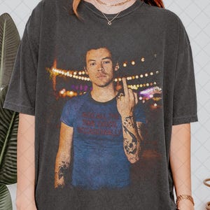 May include: Dark grey t-shirt with a portrait of a man with tattoos, giving the middle finger. The man is wearing a blue t-shirt with the text "Kiss All The Boys Occasionally". The background is blurred with lights.