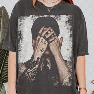 Op de afbeelding: Een antracietgrijs t-shirt met korte mouwen en een verweerde grafische print van een persoon die het gezicht met de handen bedekt. De handen tonen zichtbare tatoeages en ringen, en de persoon draagt een overhemd met kraag en das onder een vest.