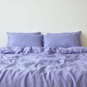 May include: A lavender-colored linen bedding set, including two pillowcases and a duvet cover, is displayed against a white wall. The fabric has a soft, wrinkled texture, suggesting comfort. The set is designed for a bed and offers a calming aesthetic.
