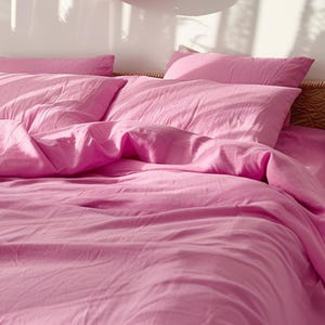 Puede incluir: Una cama con ropa de cama rosa brillante. El conjunto incluye una funda nórdica y dos almohadas. Las almohadas rectangulares están colocadas en la cama. El marco de la cama es parcialmente visible, con una textura tejida. El esquema de color es rosa liso.