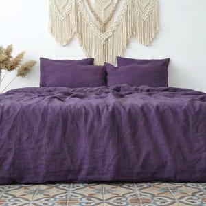 May include: A bed dressed with a deep purple linen duvet cover and matching pillowcases. The bed is made up. A decorative wall hanging is above the bed, and dried plants are to the left.