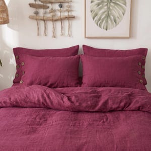 May include: A deep magenta linen bedding set consisting of a duvet cover and two rectangular pillowcases. Each pillowcase features a row of four round, light brown wooden buttons along the outer side edge, providing a textured contrast.