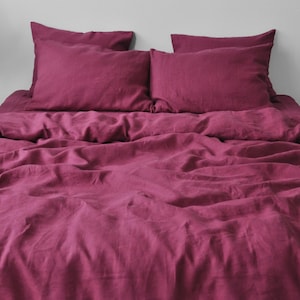 May include: A bed covered with a deep magenta-colored linen bedding set. The set includes a fitted sheet, a duvet cover, and two pillowcases. The fabric has a slightly wrinkled texture, suggesting a natural fiber.