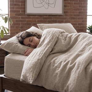 May include: A beige linen bedding set consisting of a duvet cover and two matching pillowcases. The fabric has a visible natural weave texture in a neutral light tan color. The bedding is draped over a bed frame, creating a soft and layered appearance.