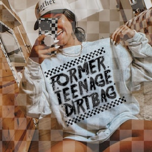 Könnte beinhalten: Hellgraues Sweatshirt mit dem Text "FORMER TEENAGE DIRT BAG" in Schwarz, umrandet von Weiß. Der Text ist von einem schwarz-weißen Schachbrettmuster umrahmt. Eine weiße Baseballkappe mit dem Wort "mother" ist ebenfalls zu sehen.