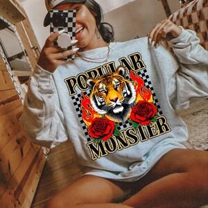 May include: A light gray sweatshirt with a graphic design featuring a tiger's face, flames, and roses. The words "POPULAR MONSTER" are printed above and below the tiger. The person in the image is wearing a white baseball cap with the word "Mother" on it.