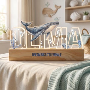 May include: A wooden night light with the name "OLIVIA" in clear letters, featuring a blue whale illustration. The base reads "DREAM BIG LITTLE WHALE". Decorative elements include bubbles, coral, a seahorse, and a seashell.