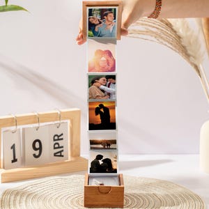 May include: A wooden photo box with a series of attached photos, held by a hand. The photos depict couples in various romantic settings. A wooden calendar displays "19 APR". A vase with dried plants is in the background.