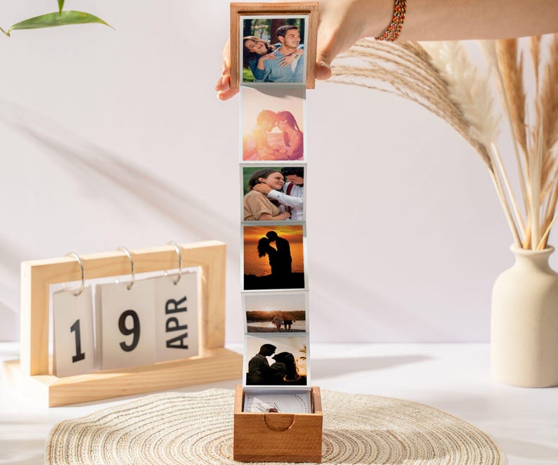 May include: A wooden photo box with a series of attached photos, held by a hand. The photos depict couples in various romantic settings. A wooden calendar displays "19 APR". A vase with dried plants is in the background.