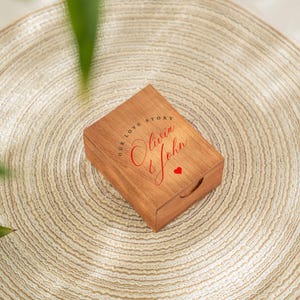 May include: A wooden box with the words "OUR LOVE STORY" and the names "Olivia & John" in red script, along with a small red heart. The box is rectangular with a handle cutout and sits on a beige woven mat.