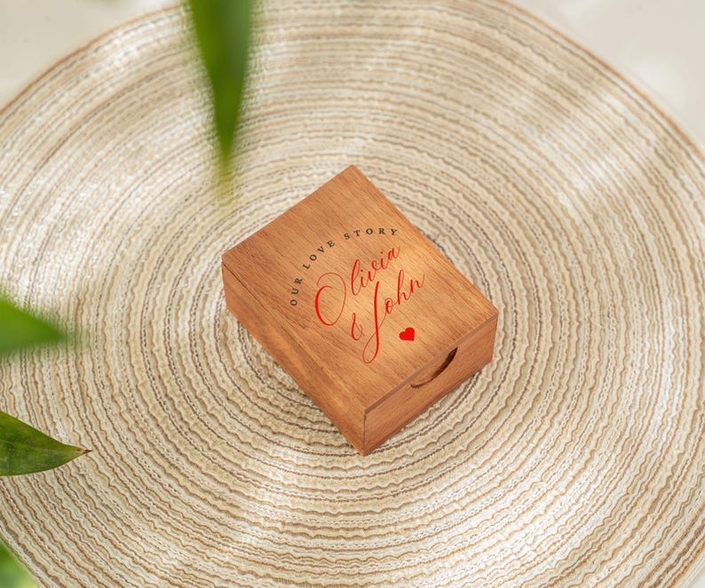 May include: A wooden box with the words "OUR LOVE STORY" and the names "Olivia & John" in red script, along with a small red heart. The box is rectangular with a handle cutout and sits on a beige woven mat.