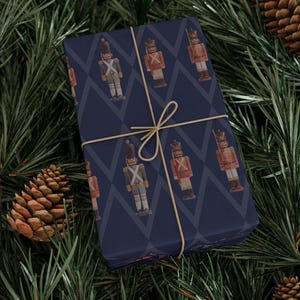 May include: A rectangular gift box wrapped in dark blue paper featuring a repeating pattern of nutcracker figures in red and blue uniforms, tied with a thin tan string. The nutcrackers are depicted in a standing position with tall hats.