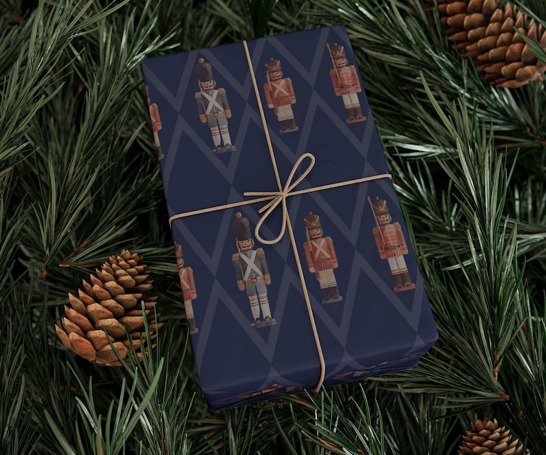 May include: A rectangular gift box wrapped in dark blue paper featuring a repeating pattern of nutcracker figures in red and blue uniforms, tied with a thin tan string. The nutcrackers are depicted in a standing position with tall hats.