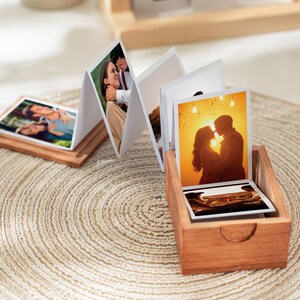 May include: A wooden photo album with a series of photographs displayed in an accordion style. The album is in a small wooden box. The photos show couples and other images. The album is on a woven mat.