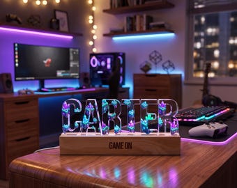 Pode incluir: Uma placa de nome personalizada em acrílico com o nome CARTER em letras de forma, apresentando um gráfico interno de cristais roxos e azuis. A placa está montada numa base retangular de madeira com o texto GAME ON impresso a preto.