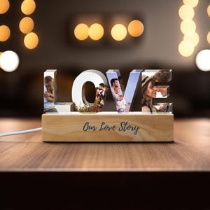 Könnte beinhalten: Ein beleuchtetes 'LOVE'-Schild mit Fotos in jedem Buchstaben, auf einem Holzsockel. Auf dem Sockel steht 'Our Love Story'. Das Schild wird von warmweißen Lichtern beleuchtet, was eine romantische Atmosphäre schafft.