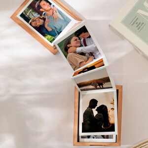 May include: A wooden photo frame with multiple pictures of couples and landscapes. The frame is made of light-colored wood and has a unique accordion-style design, displaying several photographs in a cascading arrangement. A book is visible in the background.