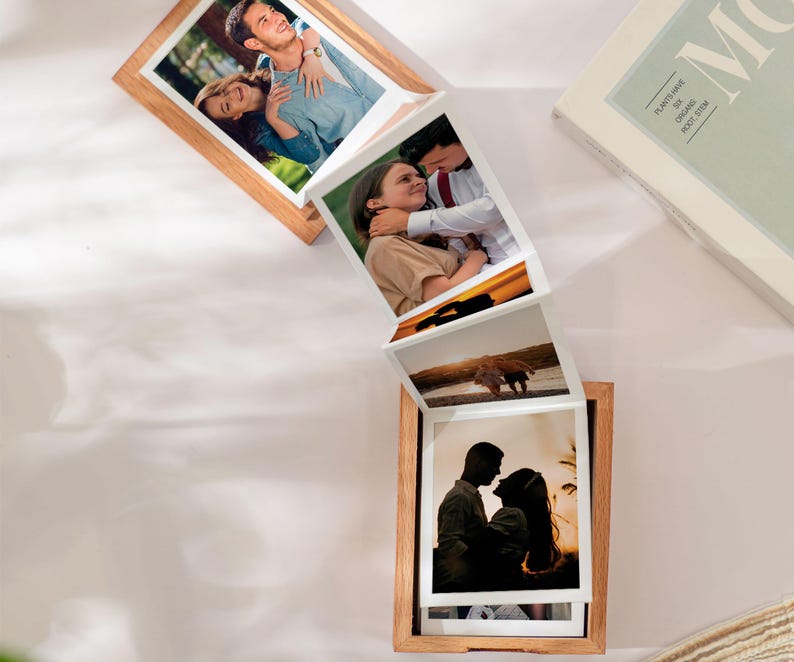 May include: A wooden photo frame with multiple pictures of couples and landscapes. The frame is made of light-colored wood and has a unique accordion-style design, displaying several photographs in a cascading arrangement. A book is visible in the background.